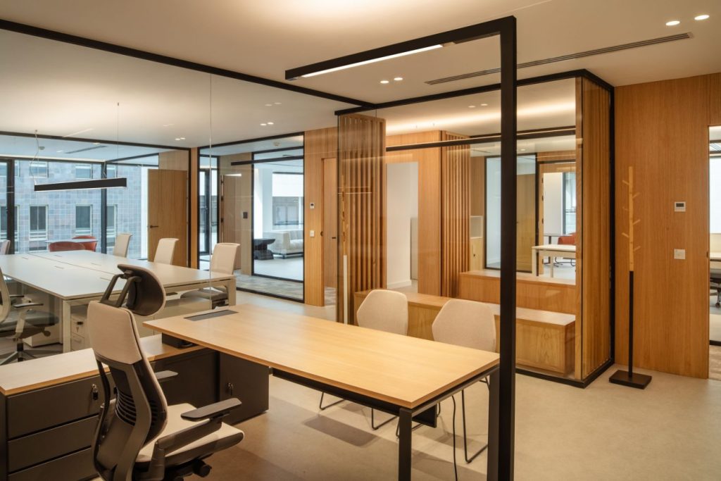 La iluminación lineal e indirecta, estratégicamente ubicada en bandejas, foseados y perfiles lineales, resalta los colores neutros, las texturas suaves y los acabados en madera de roble, creando una agradable experiencia visual de trabajo.